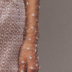 May include: A pair of sheer gloves with a delicate pattern of small white beads. The gloves are worn on a person's arm and hand.