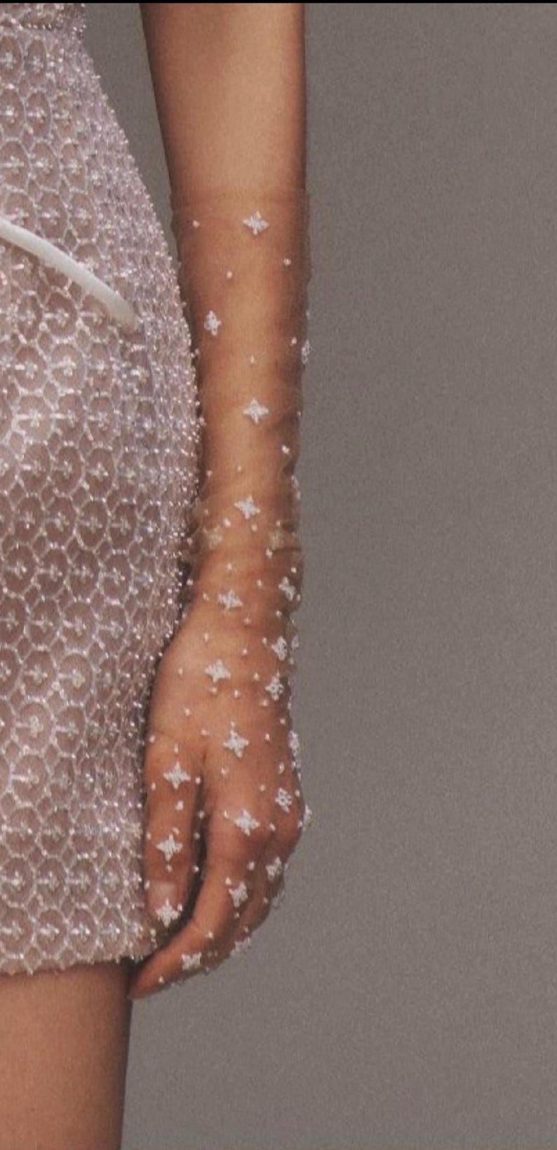 May include: A pair of sheer gloves with a delicate pattern of small white beads. The gloves are worn on a person's arm and hand.