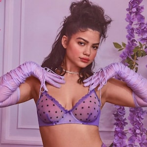 Puede incluir: Una mujer lleva un sujetador morado con estampado de estrellas negras y guantes largos a juego. El sujetador es transparente y tiene un delicado encaje. La mujer lleva el pelo recogido en una coleta alta.