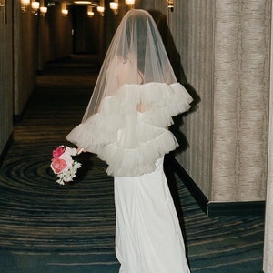 Puede incluir: Una novia con un vestido de novia blanco con un velo de tul y un ramo de flores rosas. La novia camina por un pasillo con una alfombra estampada.