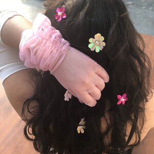May include: A pink, sheer glove with a ruffled wrist detail. Several flower hair clips in shades of pink, white, and iridescent are scattered throughout dark hair. Each flower clip has a pearl center.