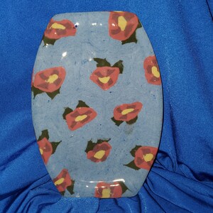 May include: Oval-shaped ceramic serving dish with a light blue background and a repeating floral pattern. The flowers are red with yellow centers and green leaves. The dish has a glossy finish.