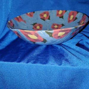May include: Hand-painted ceramic bowl with a blue background and a floral pattern. The flowers are red with yellow centers and green leaves. The bowl has an oval shape and is suitable for serving food.