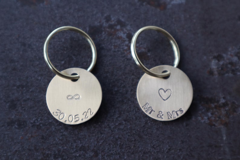 May include: Two brass keychains with round tags. One tag has an infinity symbol and the date 30.05.22 engraved on it. The other tag has a heart and the text "Mr & Mrs" engraved on it.