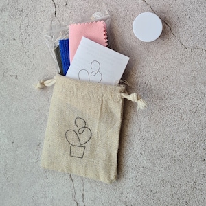 May include: A small, beige drawstring bag with a simple line drawing of a cactus plant. The bag is filled with a stack of cleaning cloths, including pink, blue, and green cloths.