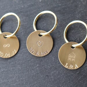 May include: Three gold keychains with round metal tags. The first tag has an infinity symbol and the date 30.05.22 engraved on it. The second tag has a heart and the text "Mr & Mrs" engraved on it. The third tag has a house and the text "L & A" engraved on it.