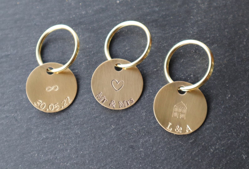 May include: Three gold keychains with round metal tags. The first tag has an infinity symbol and the date 30.05.22 engraved on it. The second tag has a heart and the text "Mr & Mrs" engraved on it. The third tag has a house and the text "L & A" engraved on it.