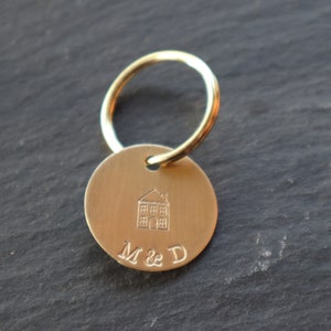 May include: Gold-toned keychain with a round tag featuring a small house illustration and the engraved text "M & D".