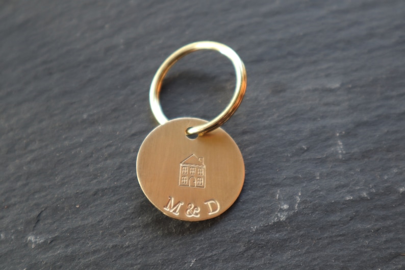 May include: Gold-toned keychain with a round tag featuring a small house illustration and the engraved text "M & D".