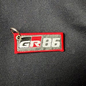 May include: A rectangular embroidered keychain featuring the text GR 86 in white and red lettering on a dark gray background. The patch is bordered by a thick red stitched edge and includes a silver metal split ring attached to the top left corner.