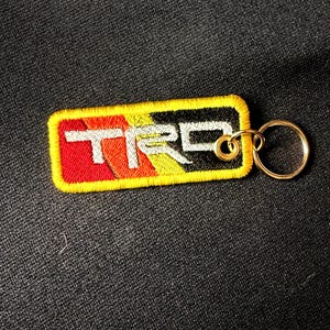 May include: A rectangular embroidered keychain featuring the letters TRD in white thread. The background consists of diagonal stripes in red, orange, and yellow, transitioning into a black section on the right. It has a yellow border and a metal ring.