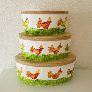 Könnte beinhalten: Drei ineinander verschachtelte Keramikschalen mit Holzdeckeln. Die Schalen sind mit einem weißen Hintergrund und bunten Comic-Hühnern verziert. Die Hühner sind gelb, orange und rot mit schwarzen Augen und Schnäbeln.