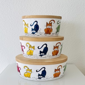 Könnte beinhalten: Drei Keramikschalen mit Holzdeckeln, jede mit einem farbenfrohen Design von Katzen mit verschiedenen Gesichtsausdrücken. Die Schalen sind übereinander gestapelt, wobei die größte Schale unten und die kleinste Schale oben ist.