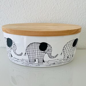 Könnte beinhalten: Weißer Keramiknapf mit Holzdeckel. Die Schüssel hat ein schwarz-weißes Elefantenmuster auf der Außenseite. Der Deckel ist aus hellem Holz. Die Schüssel steht auf einer weißen Oberfläche.