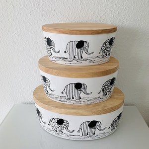 Könnte beinhalten: Drei weiße Keramik-Aufbewahrungsbehälter mit Holzdeckeln. Jeder Behälter zeigt eine Schwarzweiß-Illustration von Elefanten, die in einer Reihe laufen. Die Elefanten haben verschiedene Muster, darunter Punkte, Streifen und ein Paisley-Muster.