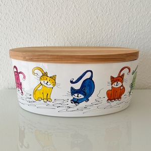Könnte beinhalten: Ein weißer Keramikbehälter mit Holzdeckel. Der Behälter ist mit bunten Cartoon-Katzen in verschiedenen Posen verziert. Die Katzen sind in den Farbtönen Rosa, Gelb, Blau, Orange und Grün gehalten. Der Behälter steht auf einer weißen Oberfläche.