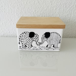 Könnte beinhalten: Eine weiße Keramik-Butterdose mit einem Holzdeckel. Die Dose zeigt eine schwarz-weiße Illustration von zwei Elefanten mit Punkten und Streifen.