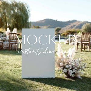 May include: A large, blank, rectangular sign with the words "MOCKUP" and "instant download" in white lettering. The sign is on a grassy lawn, with a floral arrangement and chairs in the background. The scene is outdoors.
