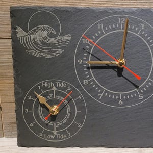 Puede incluir: Un reloj de pizarra negra con una esfera y agujas doradas. La esfera del reloj tiene los números del 1 al 12 y las agujas apuntan a las 10:50. El reloj también tiene un indicador de marea y fase lunar. El indicador de marea tiene los números del 1 al 5 y el indicador de fase lunar tiene un gráfico de la luna en diferentes fases.