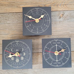 May include: Three square black slate clocks with gold-coloured hands and red second hands. One clock displays standard time, one shows moon phases with labels like New Moon and Full Moon, and one indicates tide levels with High Tide and Low Tide text.