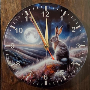 May include: A round clock featuring a white rabbit and a full moon in a snowy forest scene. The clock face has black numbers and gold hands. The clock is 30 cm in diameter.