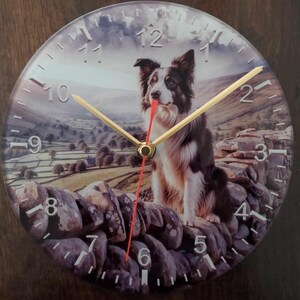 Może przedstawiać: Okrągły zegar szklany z border collie siedzącym na kamiennym murze. Tarcza zegara ma cyfry rzymskie i złotą wskazówkę godzinową i minutową. Tło to malowniczy krajobraz z zachmurzonym niebem.