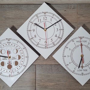 May include: Three white, diamond-shaped clocks with black hands and red second hands are displayed on a wooden surface. One clock shows the time, another displays the phases of the moon, and the third indicates high and low tide.