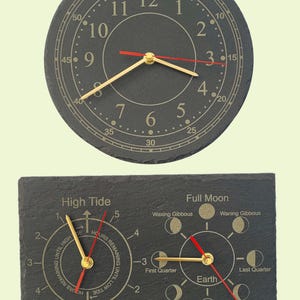 May include: A round slate wall clock with gold numerals and red and gold hands, paired with a rectangular slate tide and moon phase clock. The tide clock features gold hands and text indicating high and low tide, while the moon clock displays lunar phases.