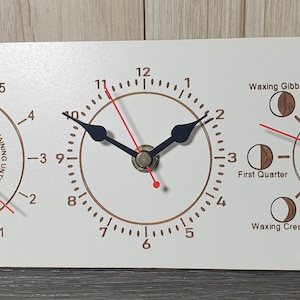 May include: A white wooden clock with three dials. The top dial shows the hours remaining until high tide. The middle dial is a standard clock face with black hands. The bottom dial shows the phases of the moon with a red hand indicating the current phase.