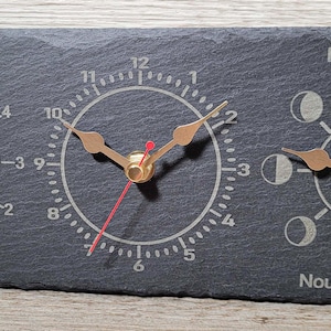 May include: A dark grey slate clock with three circular dials. The left dial indicates tide levels with the text "Marée Haute" and "Marée Basse." The centre dial shows the time. The right dial displays moon phases with the text "Pleine Lune" and "Nouvelle Lune."
