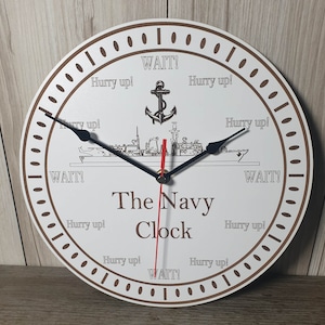 Op de afbeelding: Een witte ronde klok met bruine cijfers en een zwarte en rode wijzer. De klok heeft een marine-schipontwerp met de tekst "The Navy Clock" en de zin "Hurry up!" herhaald rond de wijzerplaat van de klok.