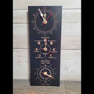 May include: A rectangular black wall clock featuring three circular dials with gold hands and red accents. The top dial displays standard time, the middle dial shows lunar phases labelled with text, and the bottom dial tracks high and low tide.