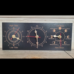 May include: A rectangular black wall clock featuring three circular dials. The left dial tracks high and low tide, the centre dial displays standard time with numbers 1 through 12, and the right dial illustrates moon phases labelled from New Moon to Full Moon.