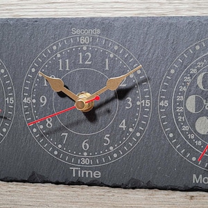 May include: A rectangular slate clock with three circular dials. The left dial displays tide information, the center dial shows the time, and the right dial indicates moon phases. Each dial has gold hands and red second hands. The clock measures approximately 30 cm long.