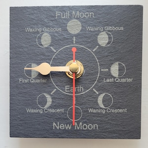 May include: A square slate wall clock featuring a lunar cycle diagram with the text Full Moon, Waxing Gibbous, Waning Gibbous, Last Quarter, Waning Crescent, New Moon, Waxing Crescent, First Quarter, and Earth. It has gold hands and a red second hand.