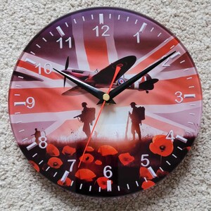 May include: A round glass clock with a red, white and blue Union Jack flag design. The clock face features a silhouette of a World War II era fighter plane, soldiers, and red poppies. The clock hands are black and red.