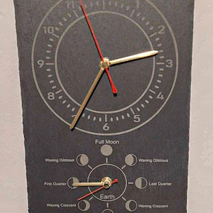 May include: A black slate clock with gold and red hands. The clock face has numbers 1 through 12 and a smaller circle with the phases of the moon. The text on the clock reads "Full Moon", "Waxing Gibbous", "First Quarter", "Waxing Crescent", "Earth", "Waning Crescent", "New Moon", "Waning Gibbous", and "Last Quarter".