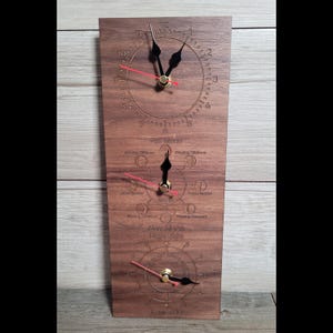May include: A rectangular wooden wall clock featuring three circular dials. The top dial displays standard time, the middle dial tracks moon phases, and the bottom dial indicates tide levels. Each dial has black hour and minute hands with a red second hand.