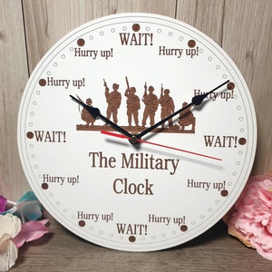 May include: A white round clock with brown text and a silhouette of soldiers. The clock face reads "WAIT!" at the top, "Hurry up!" at 3, 6, 9, and 12 o'clock, and "The Military Clock" in the center.