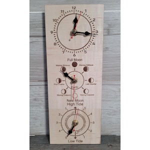 May include: A rectangular light-coloured wooden wall clock featuring three circular dials. The top dial shows standard time, the middle dial displays lunar phases labelled with moon names, and the bottom dial tracks tide levels with text indicating high and low tide.