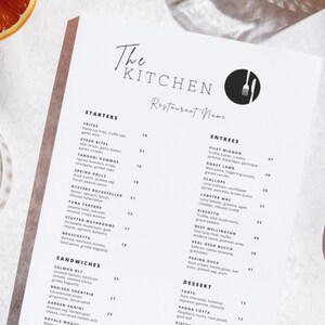 May include: A white menu with black text for The Kitchen Restaurant Name. The menu lists starters, entrees, sandwiches, and desserts. The menu is on a white surface with orange slices.
