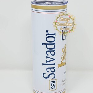 May include: A white stainless steel tumbler with a gold lid and base. The tumbler features a blue and gold logo with the text "Salvador" and a lion graphic. The tumbler also has the text "20 FL OZ" printed on the side.