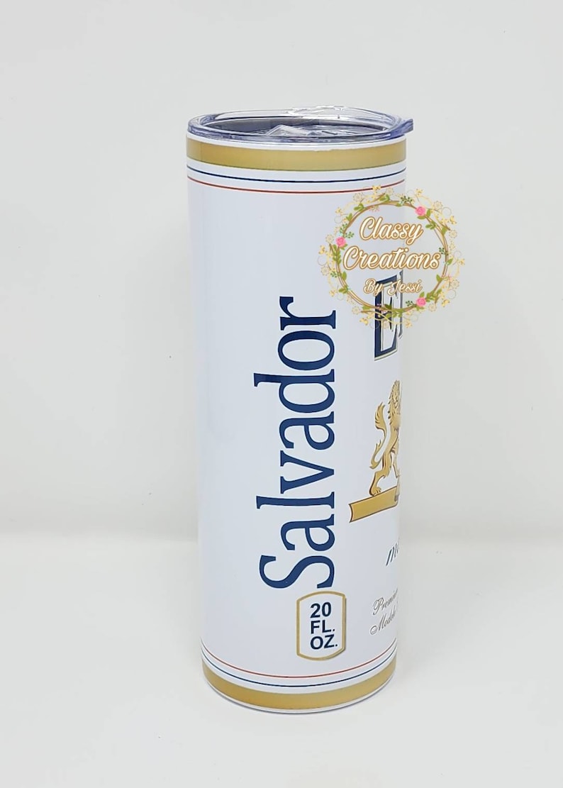 May include: A white stainless steel tumbler with a gold lid and base. The tumbler features a blue and gold logo with the text "Salvador" and a lion graphic. The tumbler also has the text "20 FL OZ" printed on the side.