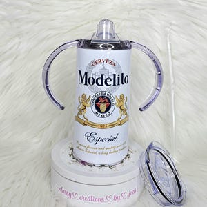 May include: A white Modelito-themed baby sippy cup with clear handles, a clear lid, and a straw. The cup features the Modelo logo and the word "Especial". The cup is sitting on a white surface.