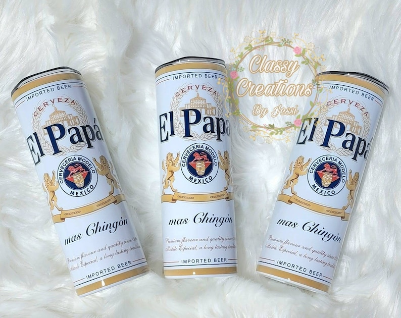 May include: Three white and blue insulated tumblers with a design that resembles a beer can. The design features the text "El Papa" and "Cerveceria Modelo" with a lion logo. The text "mas Chingon" is printed below the logo. The tumblers are on a white fuzzy surface.