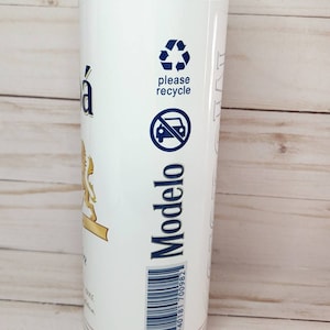 May include: A white stainless steel tumbler with a gold lid and a Modelo beer label. The label features a lion, the words "Modelo" and "please recycle" and a recycling symbol. The barcode on the label reads "40181 700982".