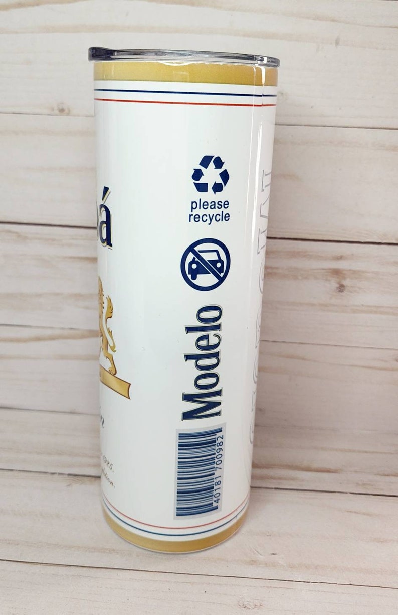 May include: A white stainless steel tumbler with a gold lid and a Modelo beer label. The label features a lion, the words "Modelo" and "please recycle" and a recycling symbol. The barcode on the label reads "40181 700982".