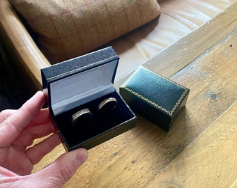 May include: Two gold wedding bands in a black velvet ring box with a green velvet ring box next to it.