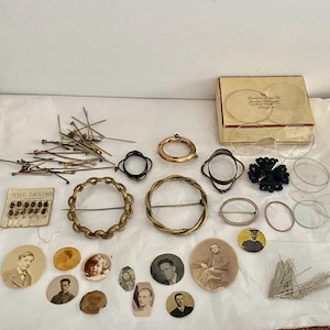 May include: A collection of vintage sewing notions and accessories including metal hat pins, circular brooches, a small card labeled The London, a box of glass lenses, a black cross-shaped ornament, and several small sepia-toned portrait photographs.