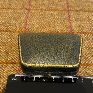 May include: A small, black, leather-bound box with a gold trim. The box is about 6 cm long.